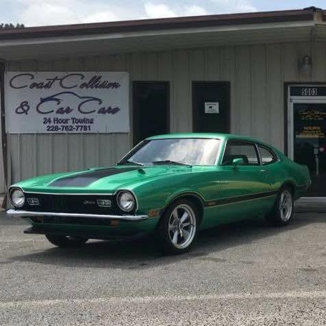 Bright green 1970s Ford Maverick with a black hood stripe parked in front of a beige metal shop building with a white Coast Collision and Car Care, 24 Hour Towing, 228-762-7781 sign
